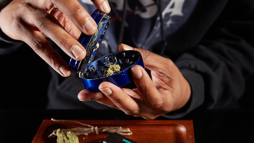 Hands opening a blue grinder filled with ground flower over a wooden rolling tray